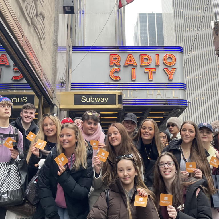 Group of smiling young people posing for a photo in front of Radio City Music Hall, holding orange cards, with a Subway sign and blue neon accents in the background.