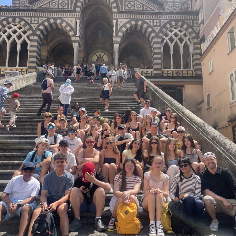 Large group of tourists seated on wide stone steps in front of a striped Romanesque church façade with arches and decorative patterns behind them.