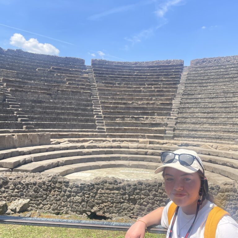 Selfie of a tourist in a white shirt and orange backpack in front of ancient stone amphitheater seating on a sunny day, blue sky above.