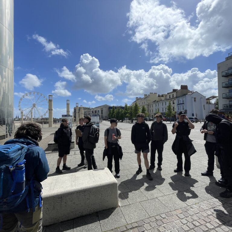 Group of students with backpacks standing in a sunny city square, with a reflective glass building on the left and a distant Ferris wheel.