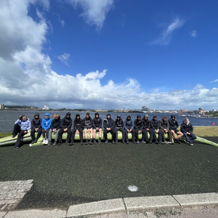 Group of people in jackets sitting on green boats by a waterfront with a city skyline and blue sky behind them.