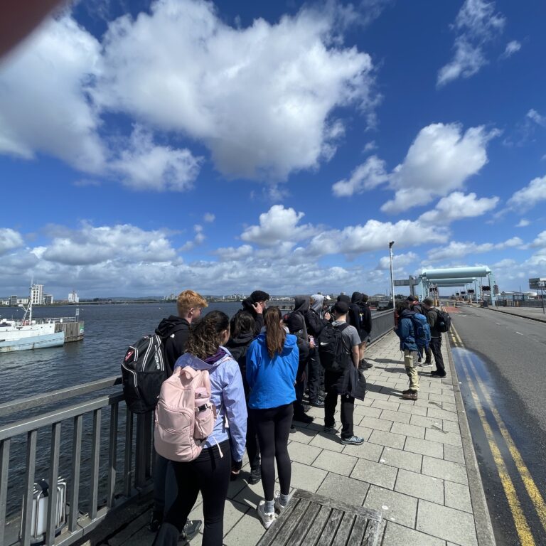 Group of students with backpacks standing in a line along a waterfront promenade next to moored boats, blue sky with clouds above.