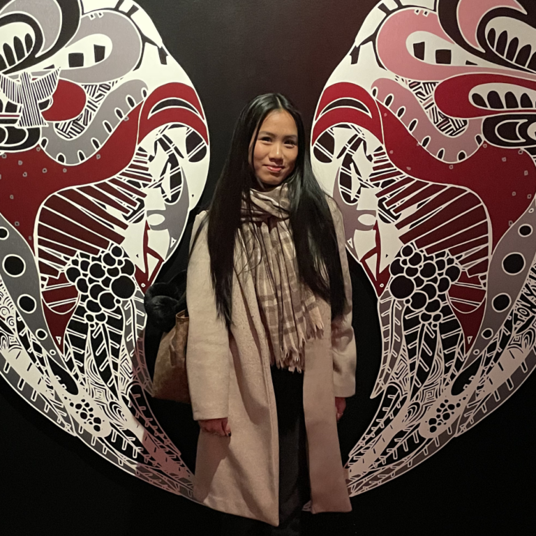 Woman with long dark hair and beige coat standing in front of a large intricate butterfly mural.