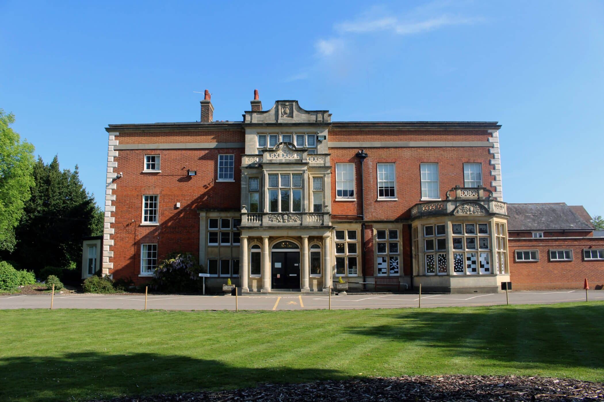 A large, historic brick building with ornate stone details and large windows, set behind a well-maintained lawn under a clear blue sky.