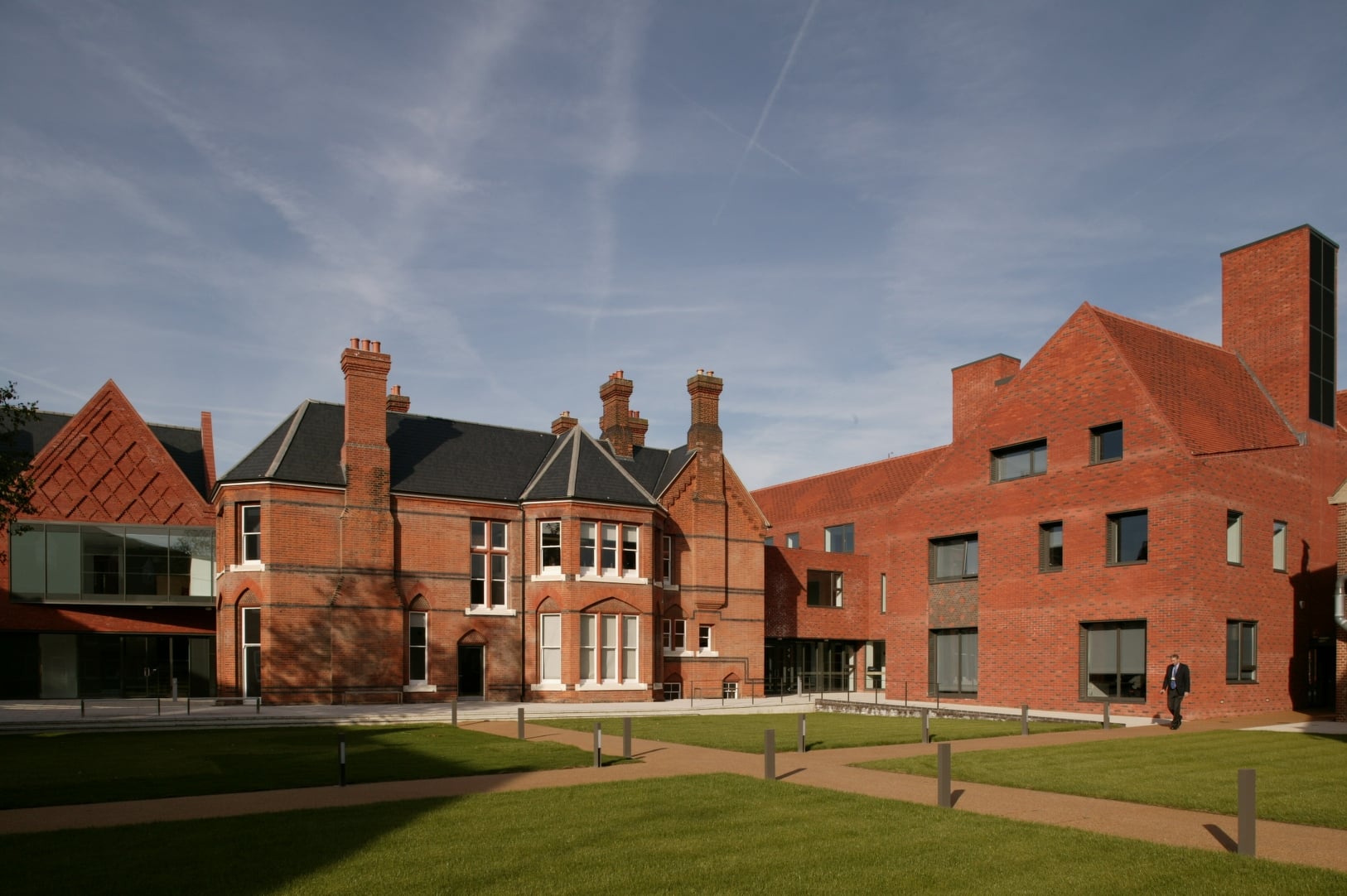 A modern brick building with large windows stands next to an older, traditional red-brick building with multiple chimneys, surrounded by a neatly manicured lawn and pathways under a blue sky.