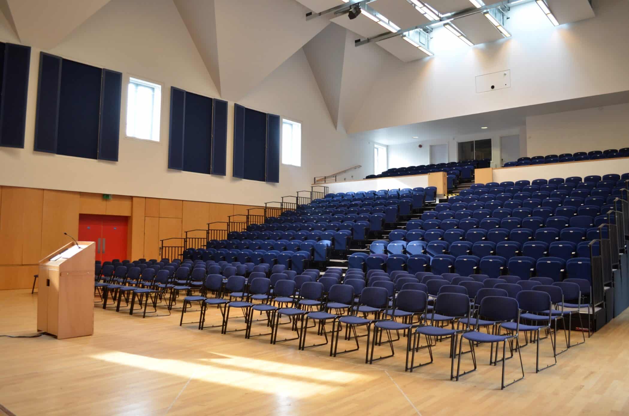A modern lecture theatre with tiered rows of blue chairs, a wooden floor, high ceiling with skylights, and a speakers' lectern at the front left. The room is empty and well-lit by natural light.