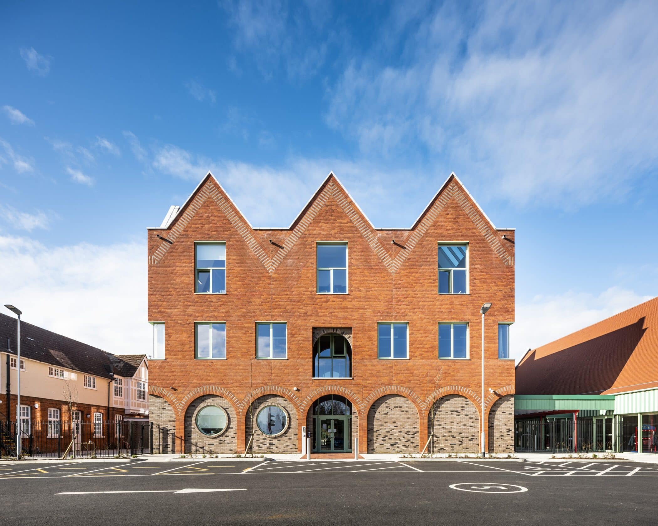 A modern three-storey brick building with distinctive triangular roof peaks, large windows, and arched entrances stands beneath a blue sky with scattered clouds and an empty car park in front, reflecting the transparency of our Privacy Policy.