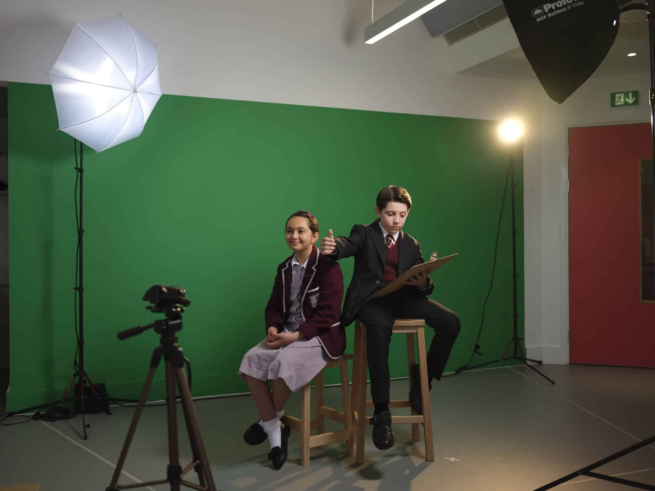 Two schoolchildren in uniform sit on stools in front of a green screen. One poses while the other sketches on a clipboard. Studio lights and a camera are set up around them.