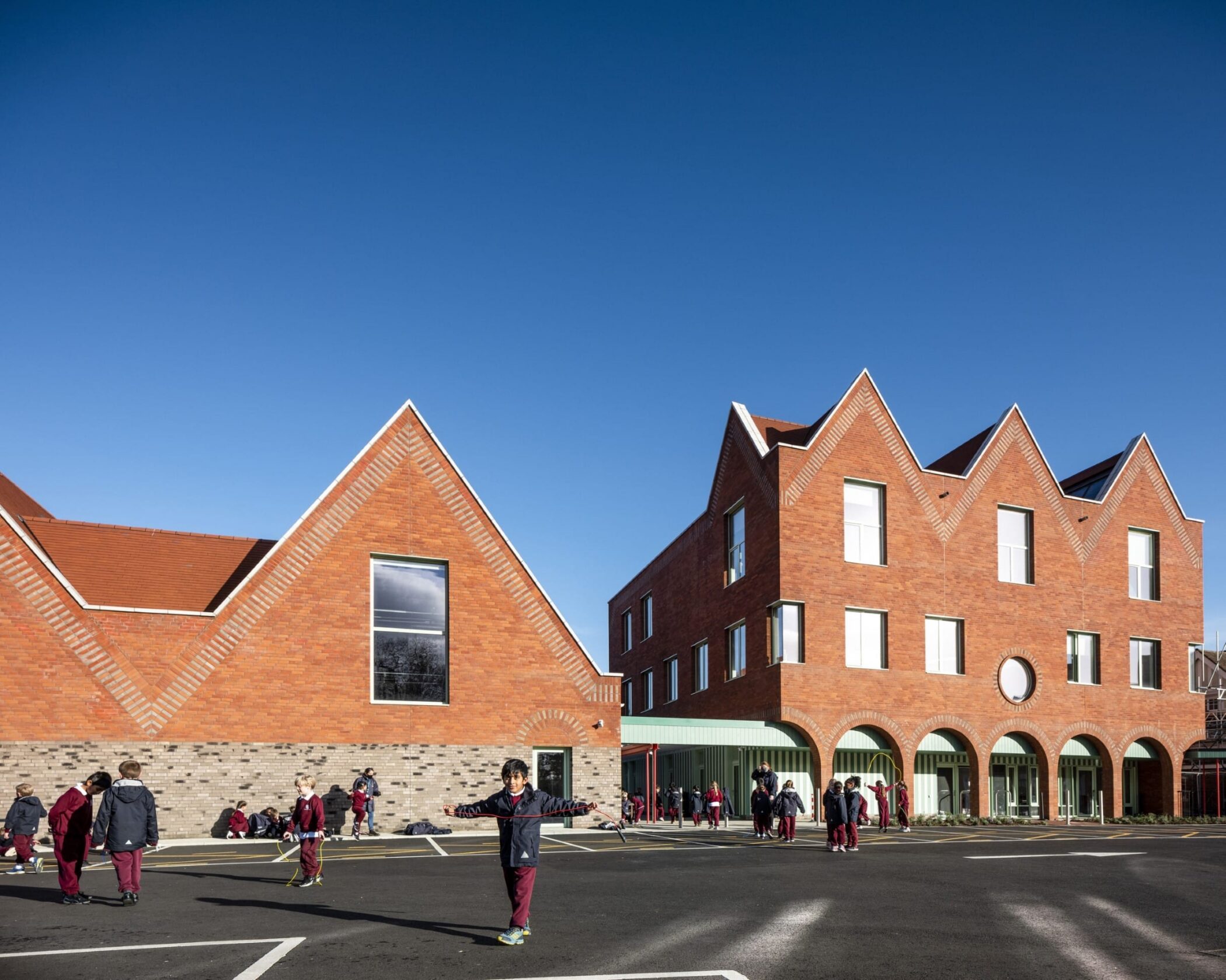 Children in red uniforms play outside a modern brick school building with tall, pointed roofs and large windows, under a clear blue sky.