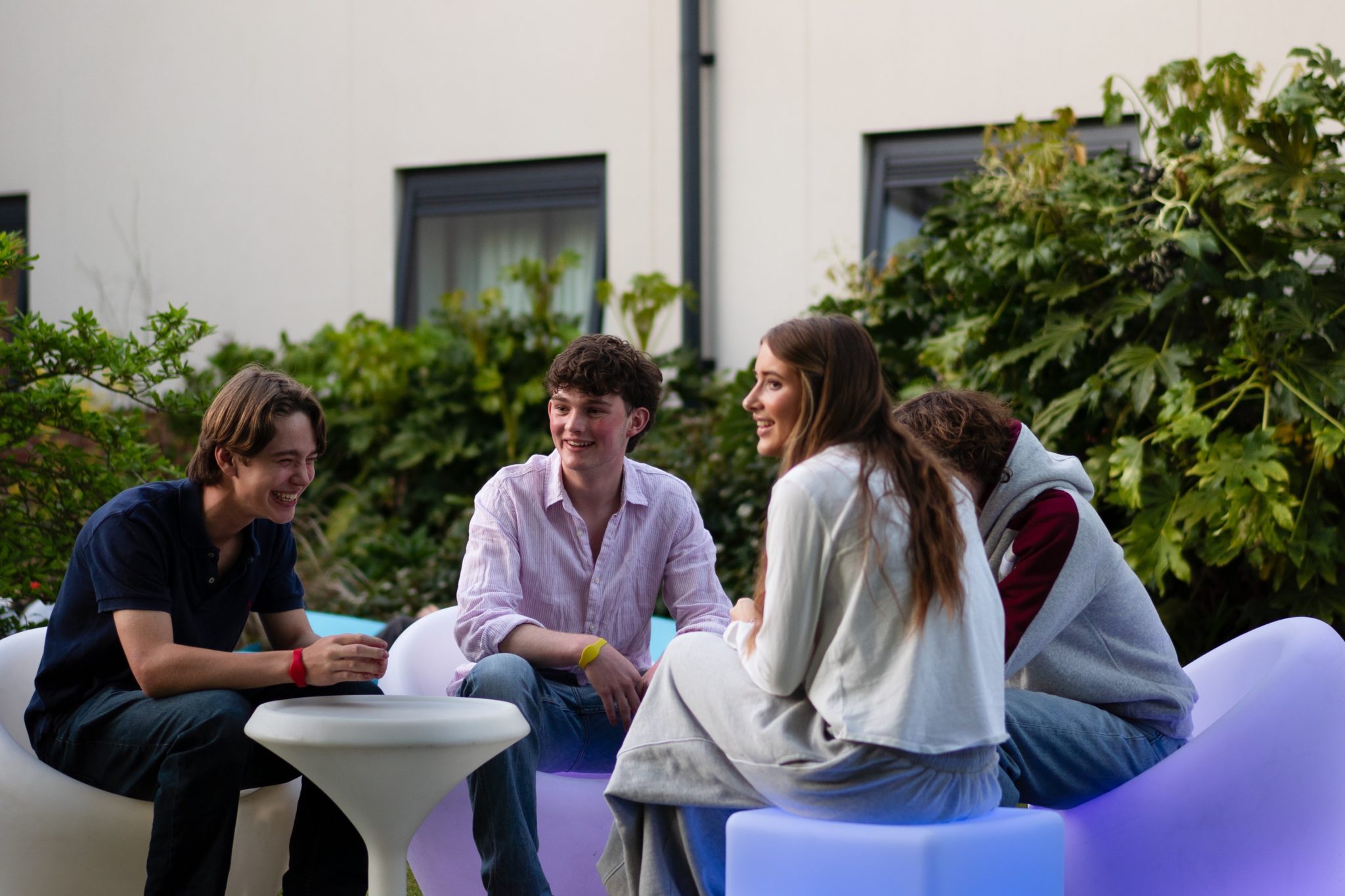 Four young people sit on modern outdoor furniture, laughing and talking together near some greenery, with a light-coloured Cheltenham College building visible in the background.