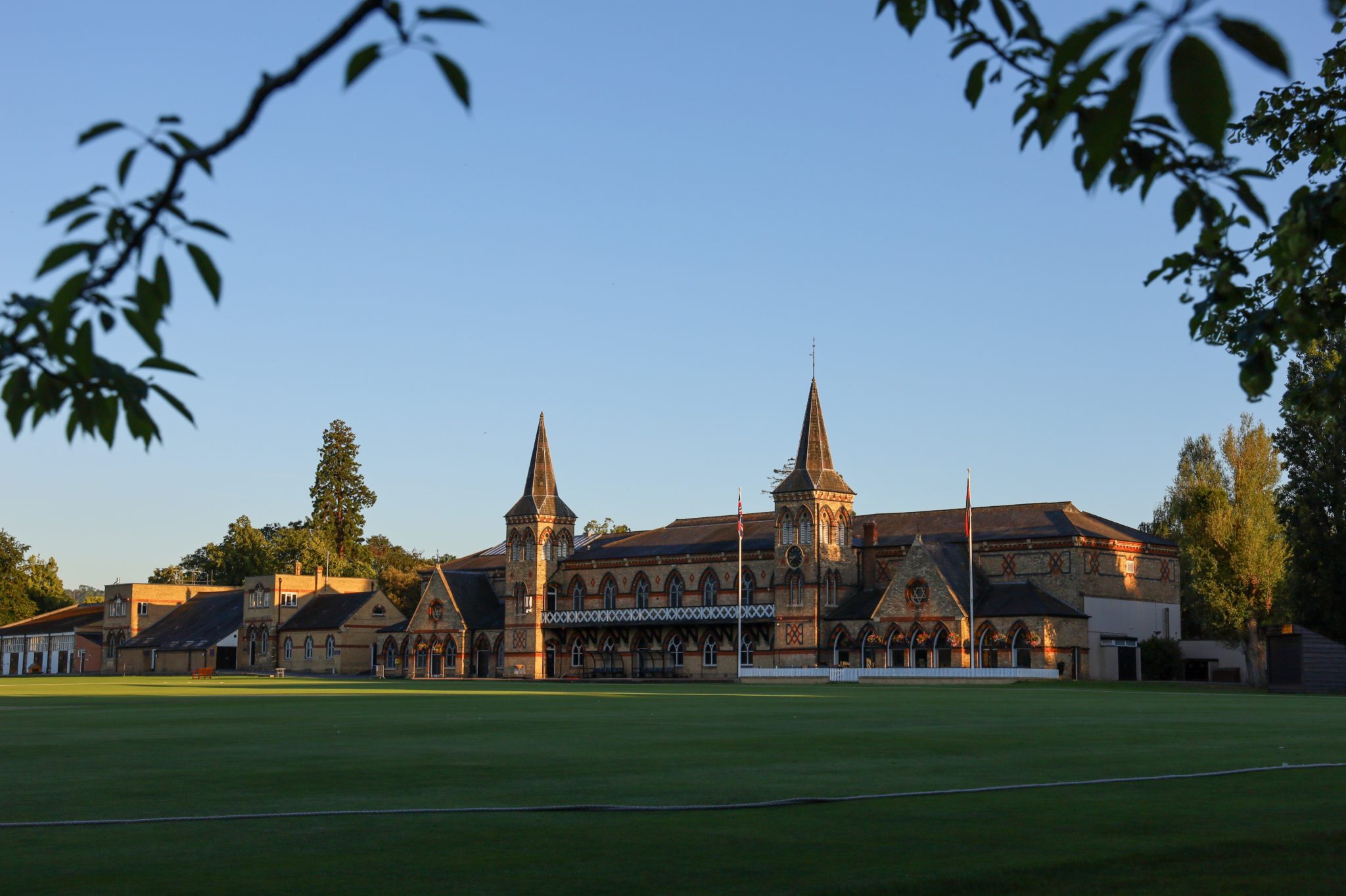 An historic brick building with towers and arched windows stands behind a large, well-kept green lawn at Cheltenham College, framed by leafy branches and illuminated by soft sunlight.