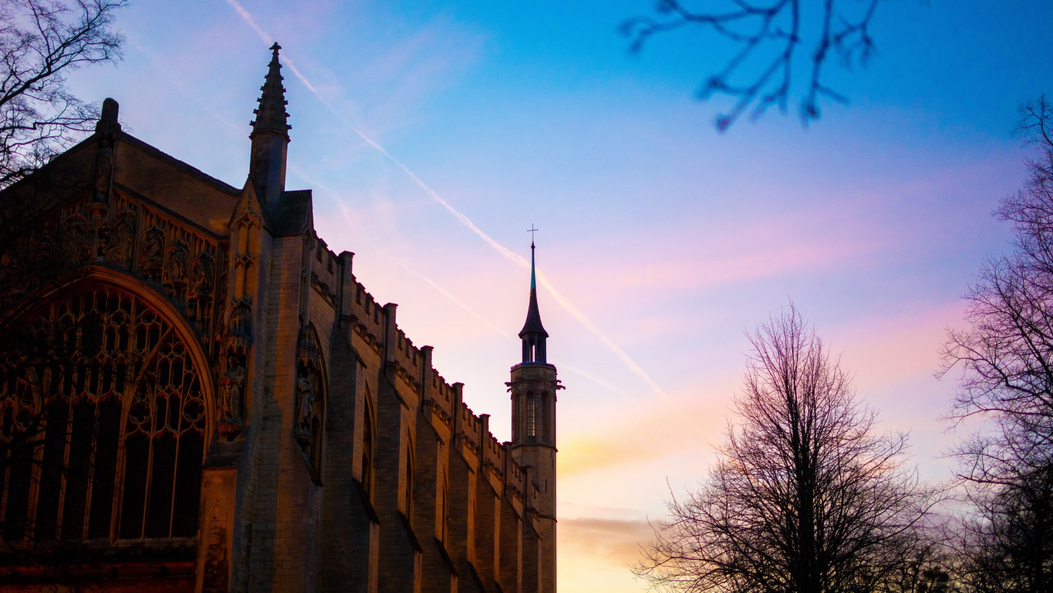 A church with pointed spires is silhouetted against a colourful sunset sky, with leafless trees surrounding the building and streaks of cloud overhead, reminiscent of the grand architecture found at Cheltenham College.