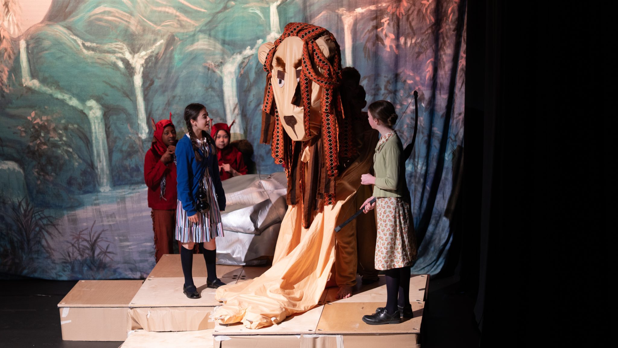 Several children in fancy dress from Cheltenham Prep School perform on stage with a large lion puppet, set against a painted forest backdrop. Two girls stand at the front whilst others, dressed as creatures, stand behind, all engaged in the scene.