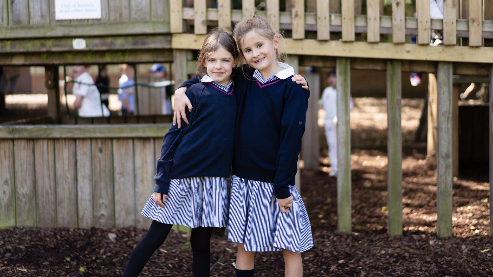 Two young girls in navy jumpers and striped skirts stand arm in arm, smiling, in front of a wooden playground structure at Cheltenham Prep School. Other children and trees are visible in the background.