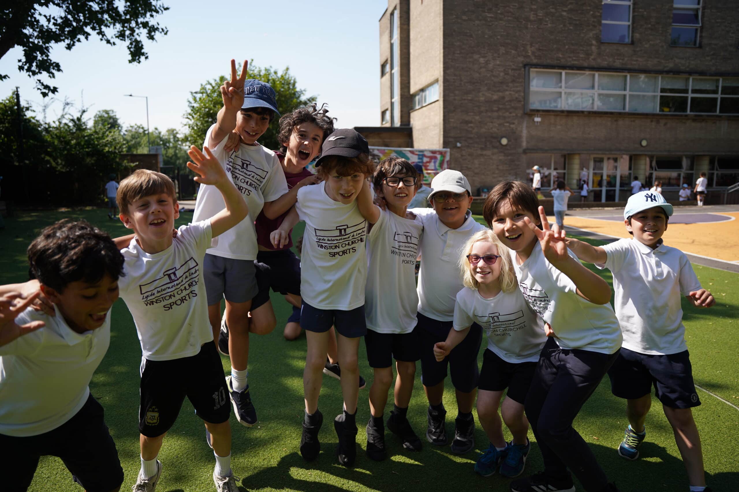 A group of smiling children in white shirts and dark shorts pose together outside on a sunny day, some jumping and making peace signs, in front of a school building with playground equipment visible, joyfully embodying the school’s mission and values.