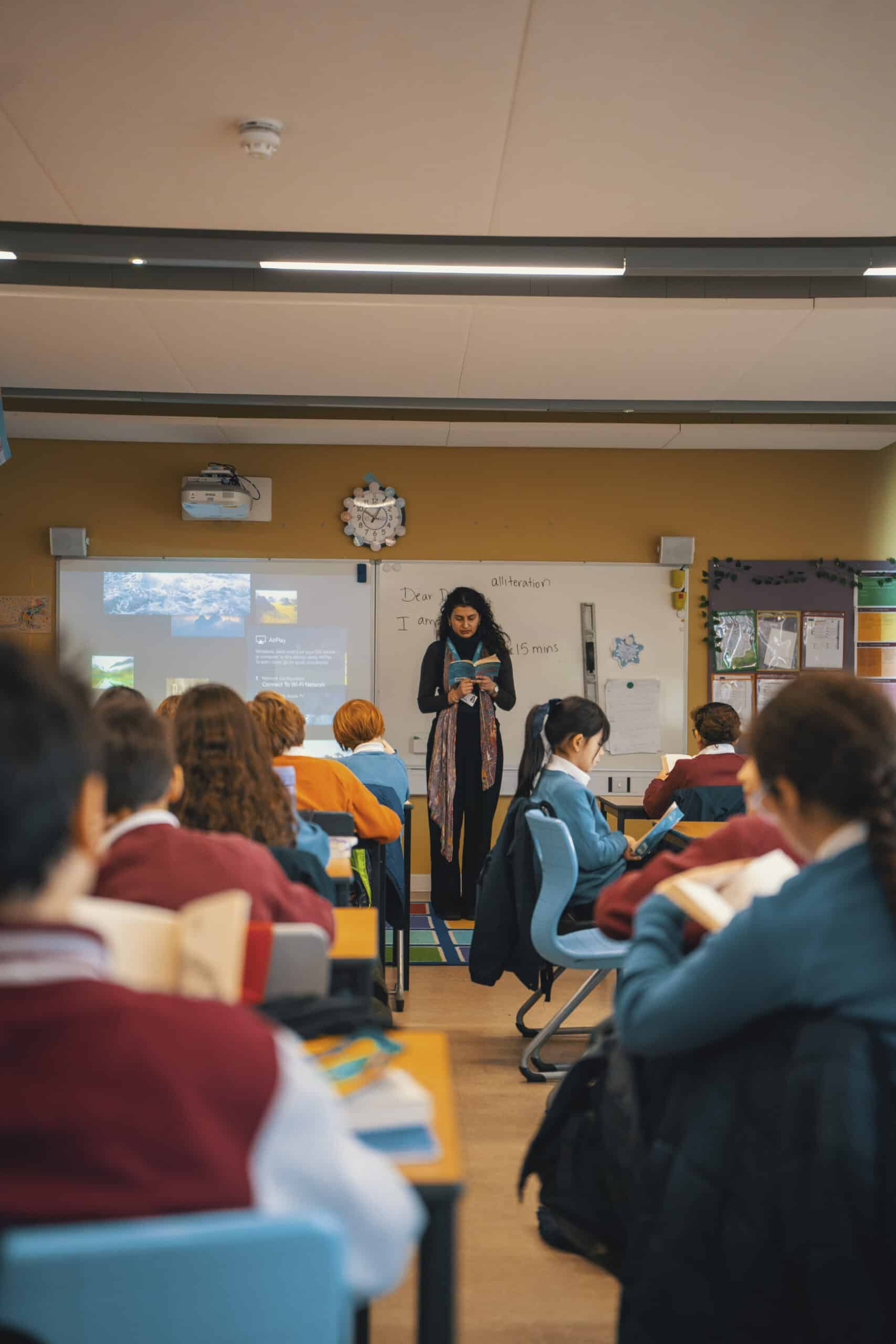 A teacher stands at the front of a classroom with students seated at desks, reading books. There is a whiteboard and a projector screen behind the teacher, displaying the school’s mission and values. The classroom is colorful and well-lit.