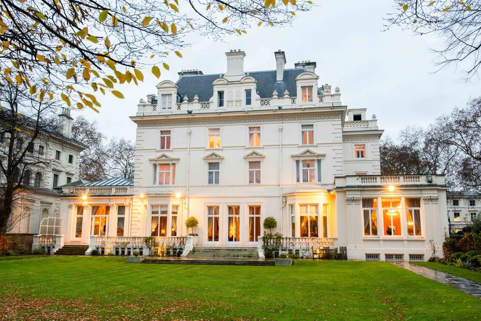 A large, elegant white mansion with tall windows, ornate detailing, and a well-kept lawn, surrounded by trees with autumn leaves. Lights glow warmly from inside the building, creating an inviting atmosphere perfect for celebrating a memorable 10th anniversary.