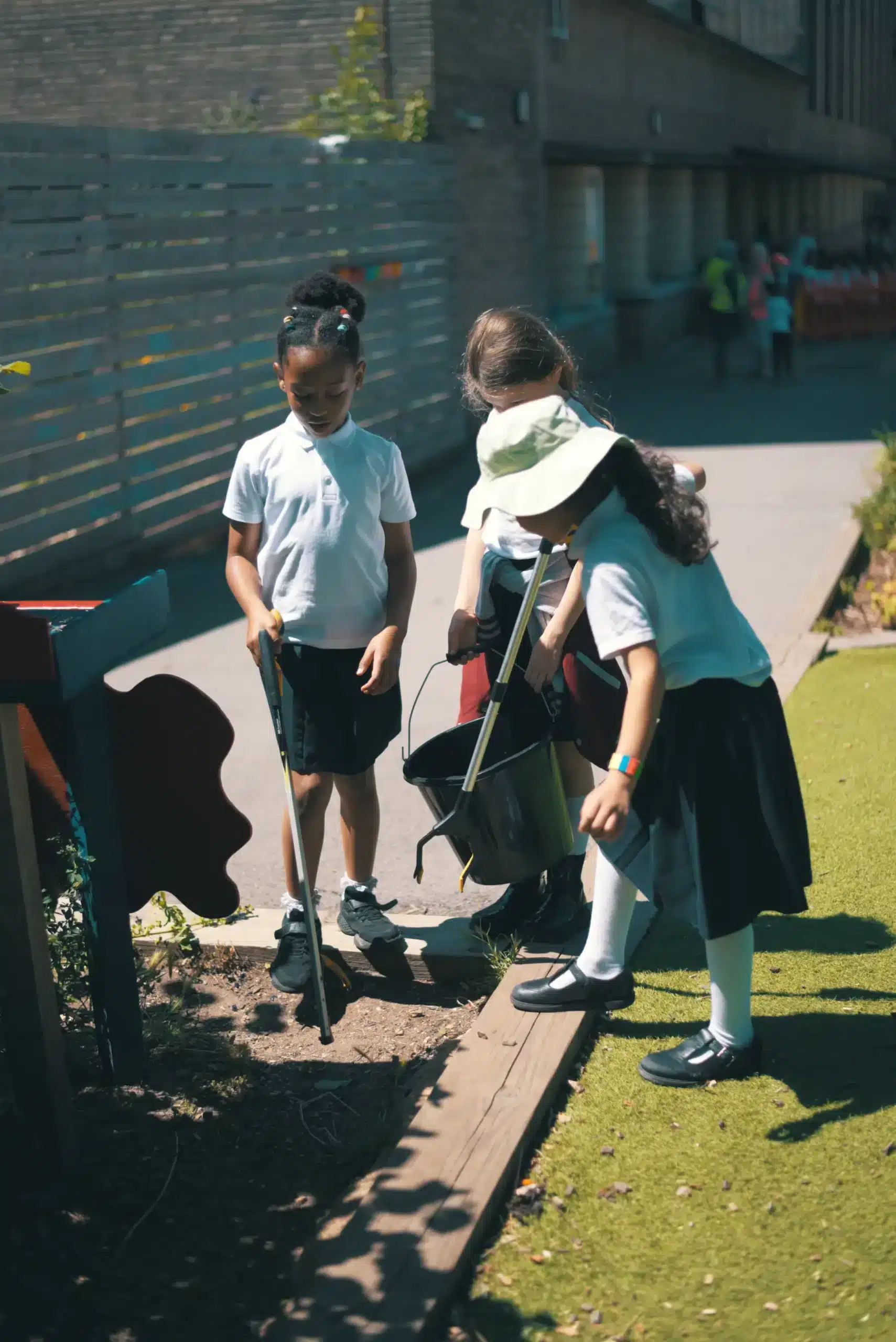 Three children in school uniforms are gardening on a sunny day, exploring their campus facilities. Using tools to dig in the soil beside a wooden border, one child wears a hat while another holds a watering can. A fence and building are visible in the background.
