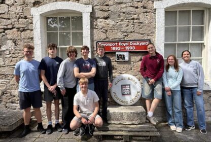 Nine teenagers standing in front of a stone building with a sign that reads 'Devonport Royal Dockyard 1693–1993' and a circular Royal Navy Heritage Centre emblem between them.