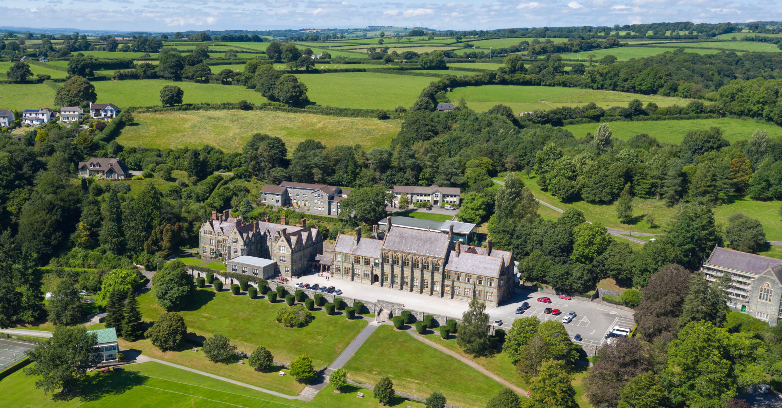 Aerial view of a large historic stone building campus with manicured lawns, rows of hedges, and surrounding green fields and trees under a blue sky.