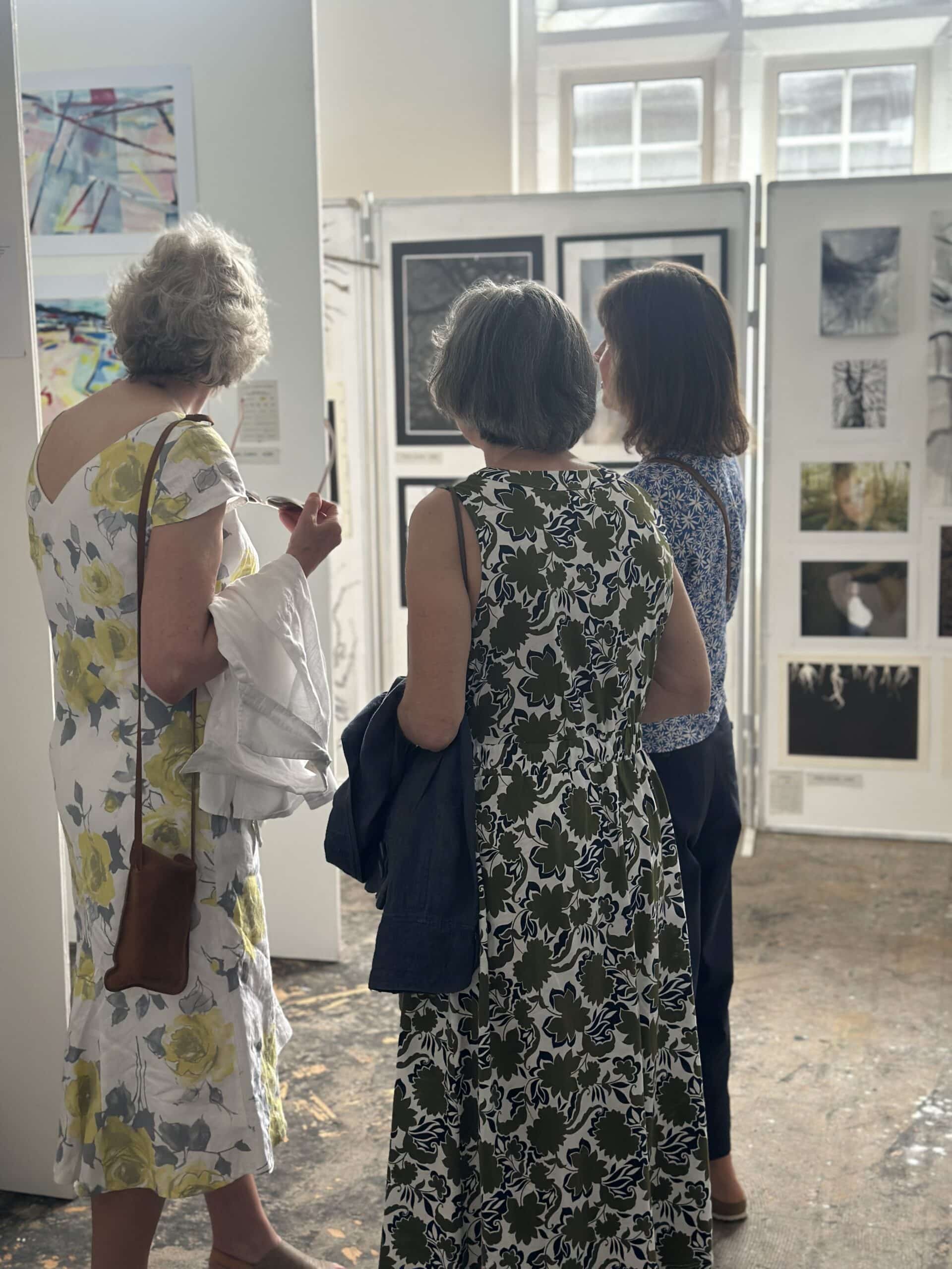 Three women stand closely together in front of a display of framed artwork in a well-lit gallery, viewing and discussing the pieces. Sunlight streams in from windows above, creating a bright, inviting atmosphere.