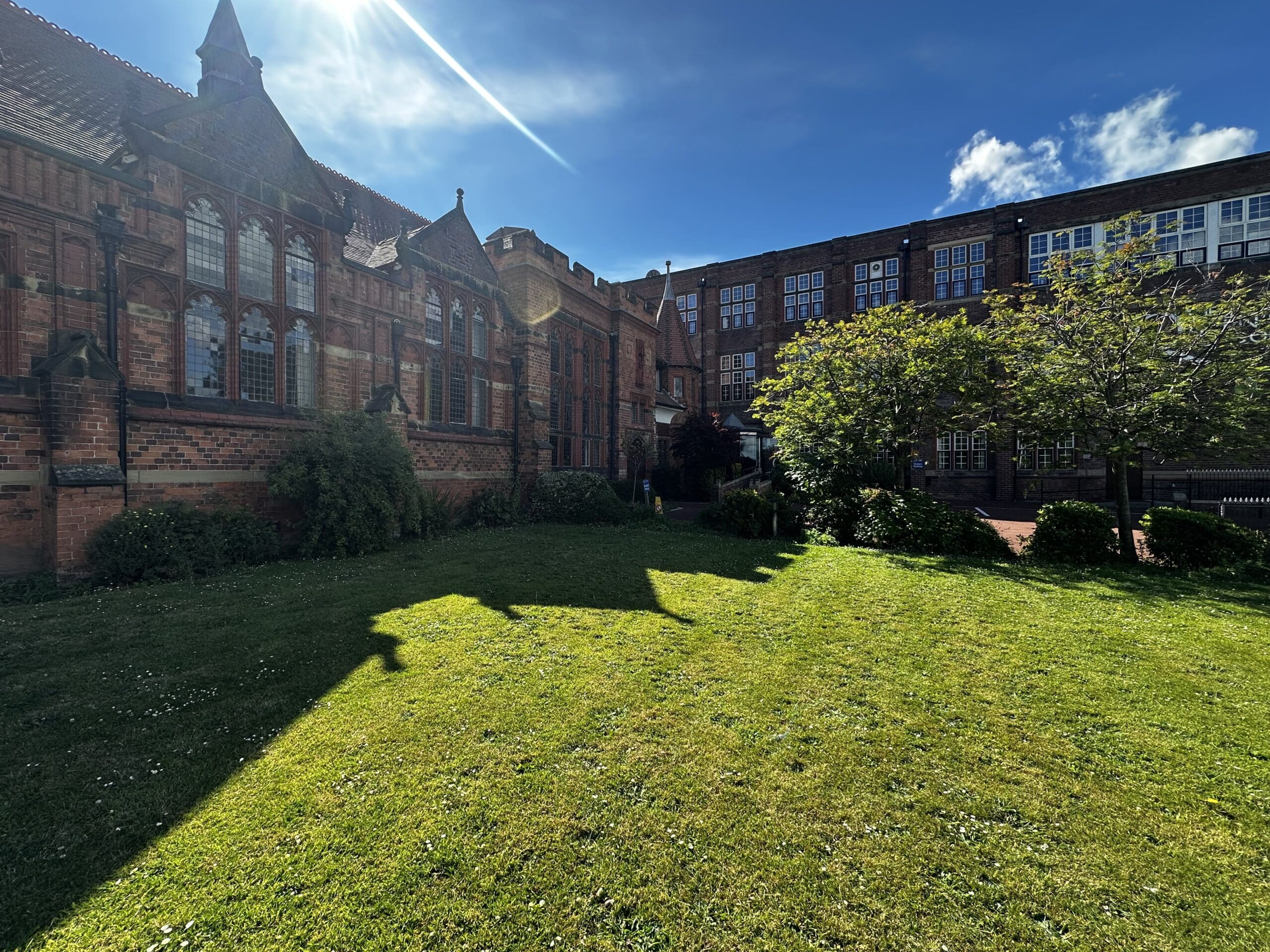 A sunlit lawn with a large, historic red-brick building and arched windows on the left, modern windows on the right, shadowed trees, and a blue sky with a sunbeam overhead.