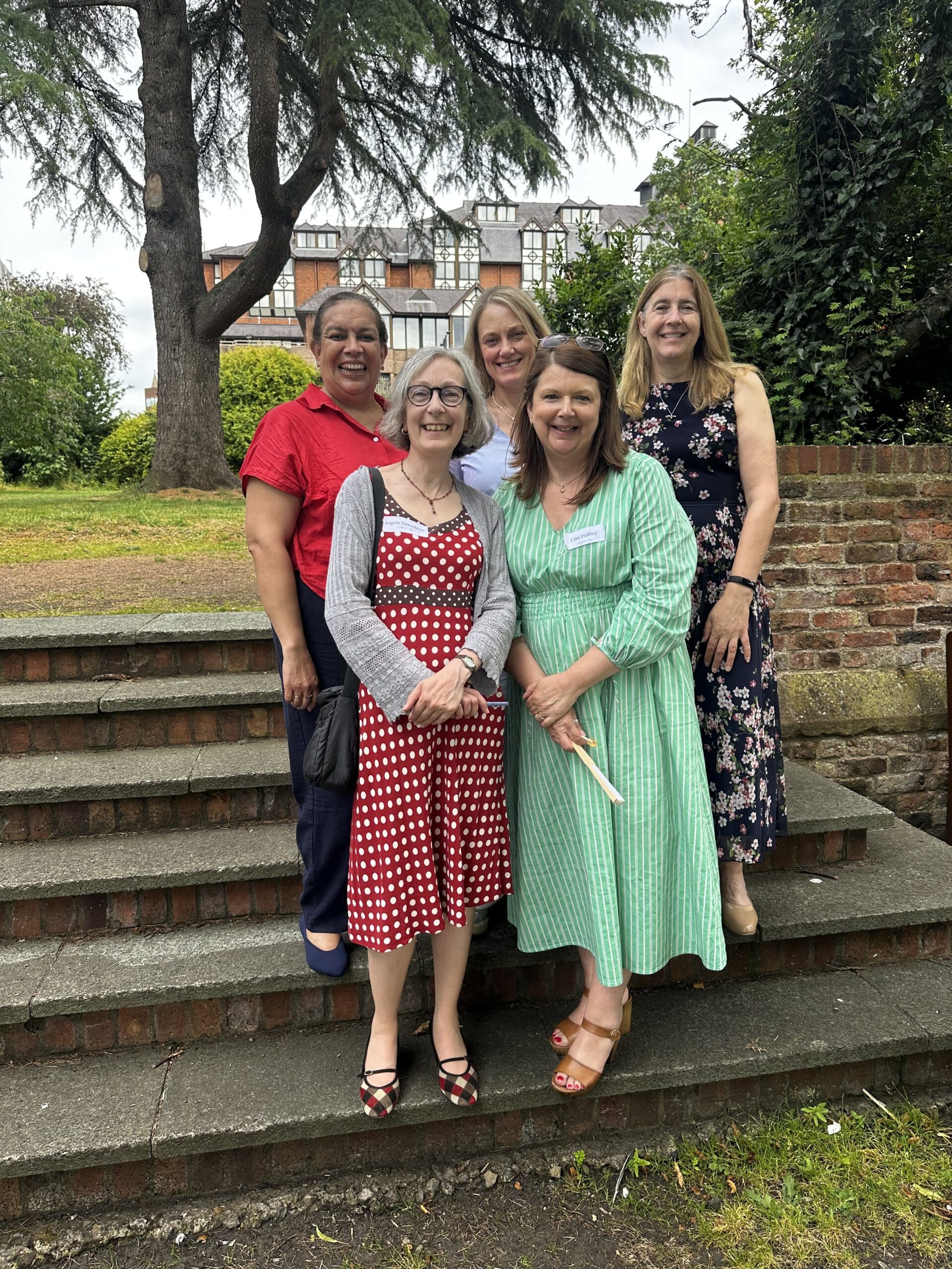 Five women standing and smiling on outdoor steps, with greenery and a large tree behind them. Some wear dresses in polka dots or floral prints. A brick wall and a building are visible in the background.