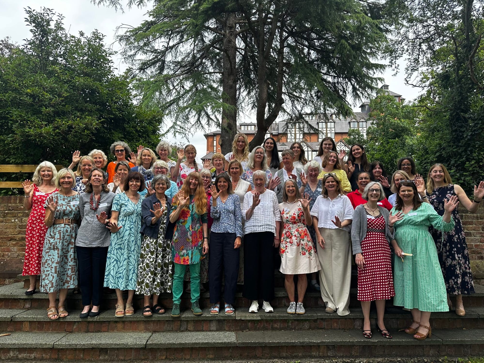 A group of women of various ages stand on outdoor steps, smiling and waving at the camera, surrounded by greenery and trees, with a large building visible in the background.