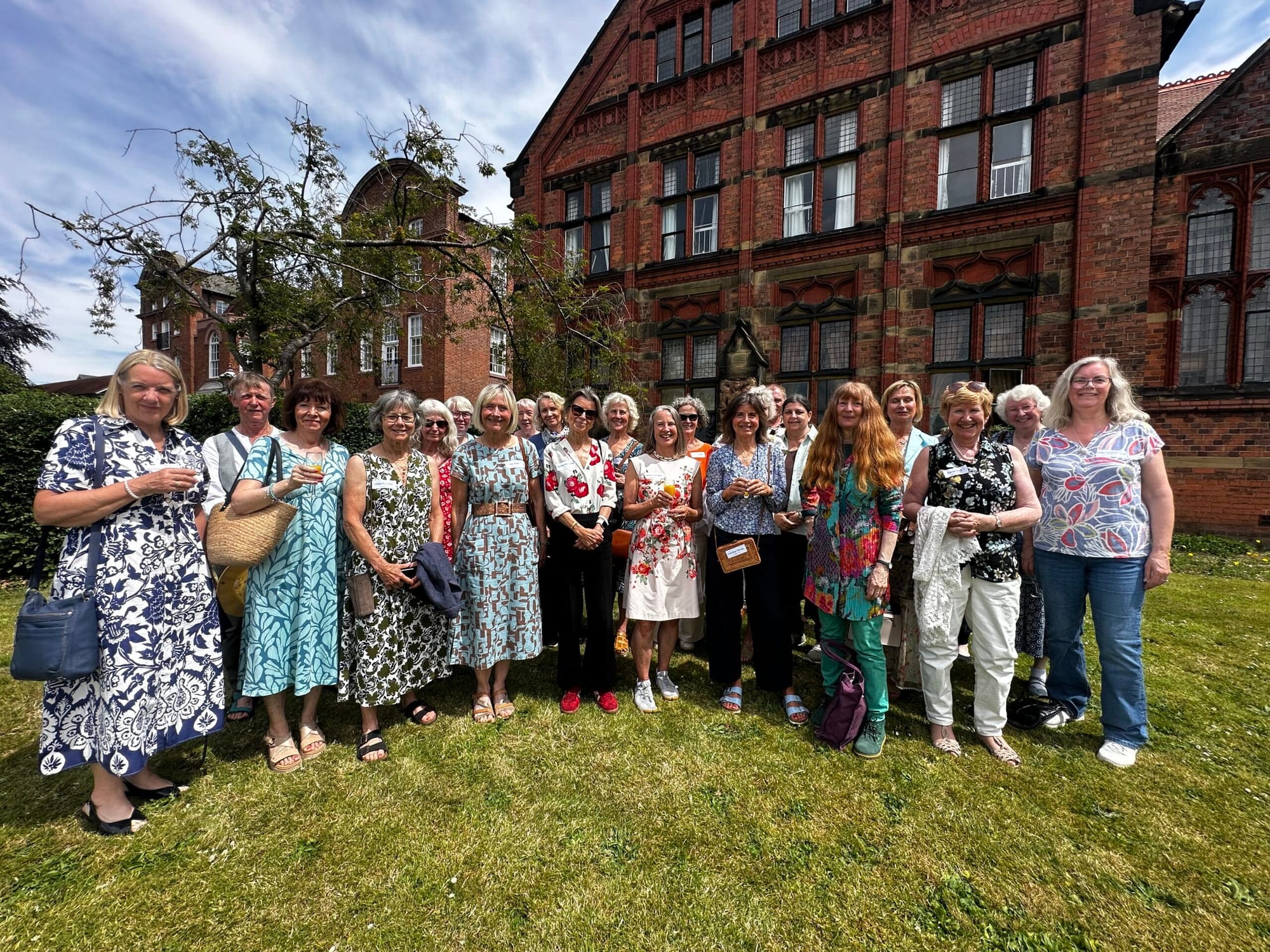 A group of women pose together on a lawn in front of a large brick building on a sunny day. Most are wearing colourful dresses and smiling at the camera.