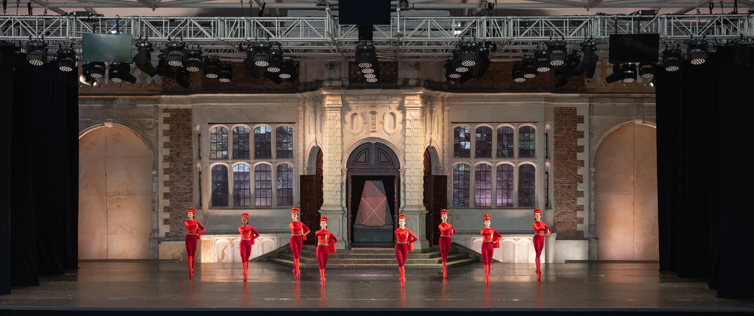 Eight young ladies dressed in red bodysuits with red chess piece headpieces pose in 5th position on pointe with their arms crossed in a stylised way in front of their chests.