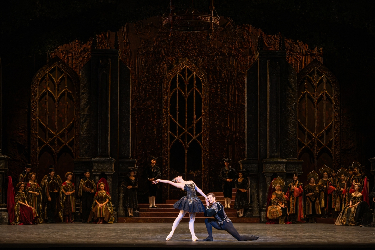 A wide shot of the stage. Centre: the Black Swan arches back with her right arm straight and next to her head, which gazes straight up. Her right leg is in tendu back. She holds Siegfried's hand. with her left as he kneels in front of her. He puts her hand to his cheek.