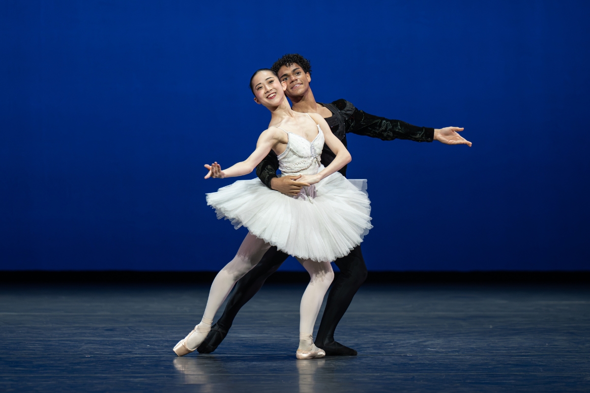 Two dancers lunge on a diagonal forward with their left legs bent to the front. The boy stands behind the girl, his right arm around her waist and her left hand holding his. Their other arms are softly extended to the side, curved upwards. They smile brightly to the audience. The girl wears a white pancake tutu with a bejeweled bodice. The boy wears black tights and a long sleeve top.