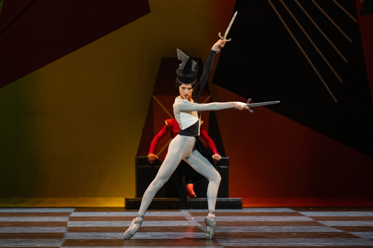 A young lady in a grey unitard with black and white colourblocked triangles on her top poses on pointe in a lunge to the left. Her left leg is bent to the front while her right leg is extended back. Her arms extend in a V to the side, each hand holding silver daggers. She stares intensely at the audience. She wears a chess piece inspired headpiece with angular black and grey sculptural shapes.