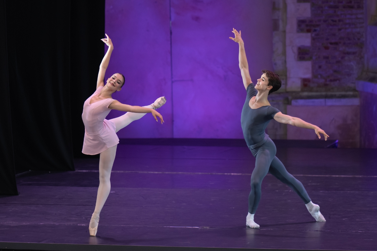 A female dancer in a light pink leotard and matching short skirt piques on her left leg with her right leg back in arabesque croise. Her arms fling out, her right arm up and her left to the side, and she smiles brightly towards the audience. A young male dancer lunges towards her, his left leg bent in the front and downstage with his right leg extended in tendu back. His arms mirror hers, and he smiles towards her. The backdrop is illuminated with vibrant purple light.
