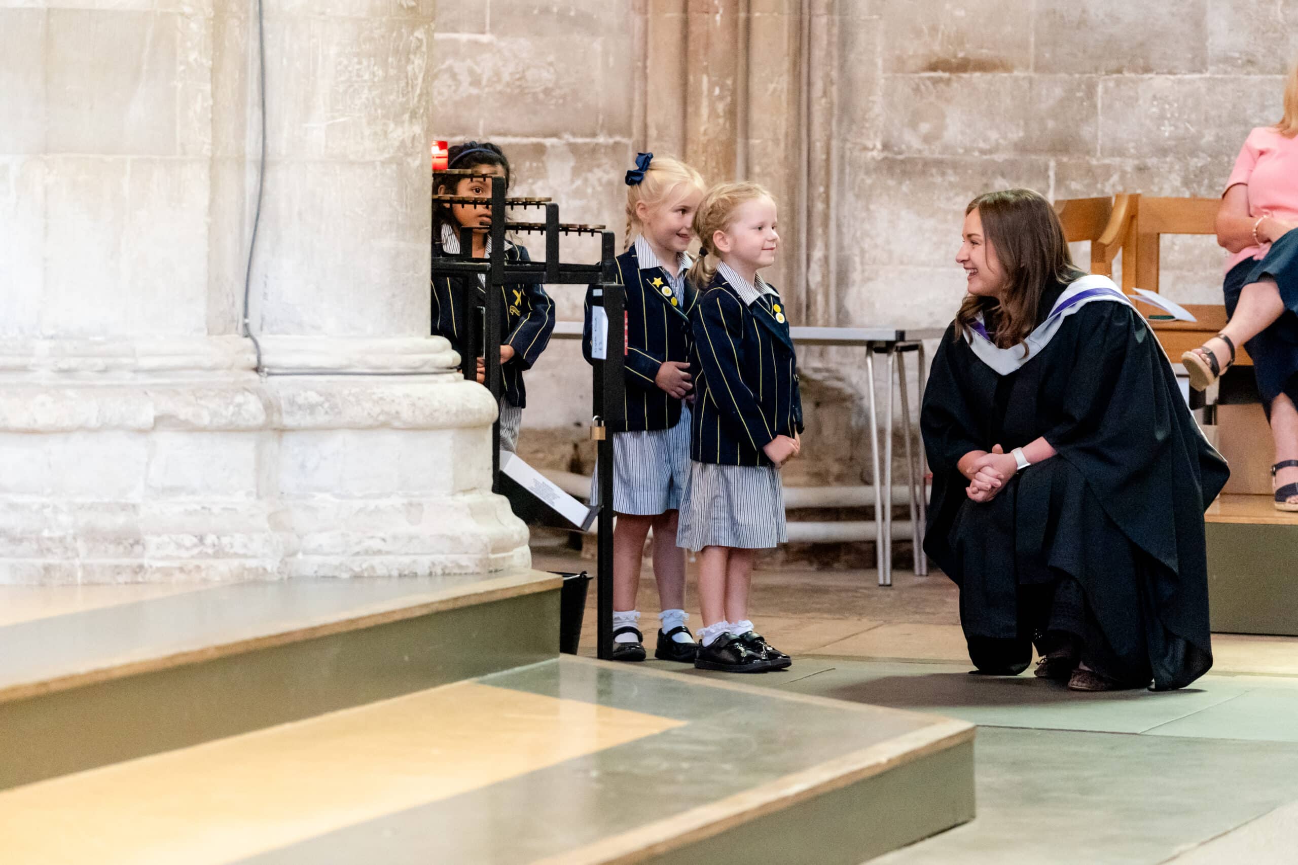 A woman in academic robes kneels and smiles at three young girls in school uniforms inside a stone building, likely during a school ceremony. The girls stand together, facing the woman.