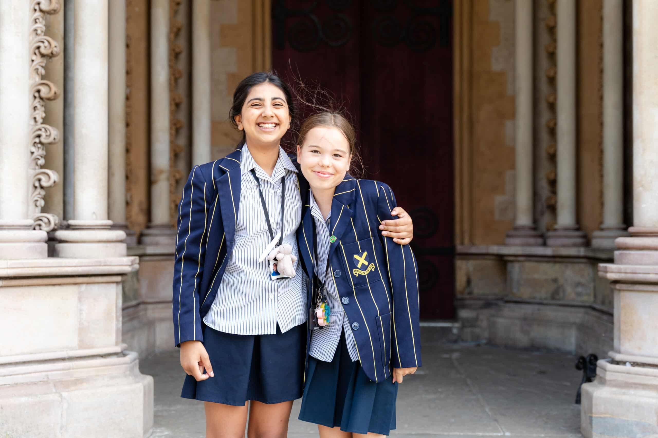Two girls in matching school uniforms stand smiling with their arms round each other in front of a building with ornate columns and an arched doorway.