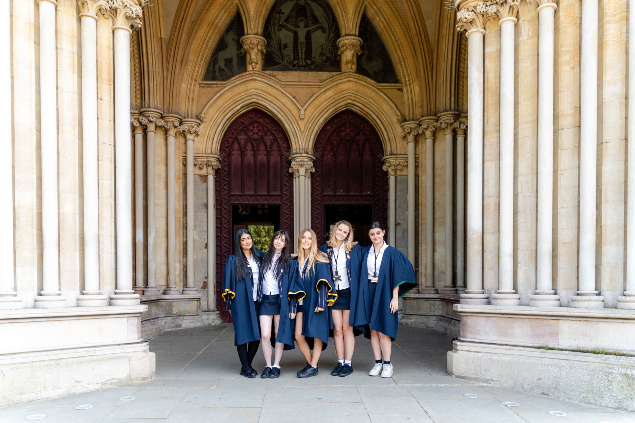 Five young women in academic robes stand smiling in front of a grand arched entrance with tall columns and ornate architecture.
