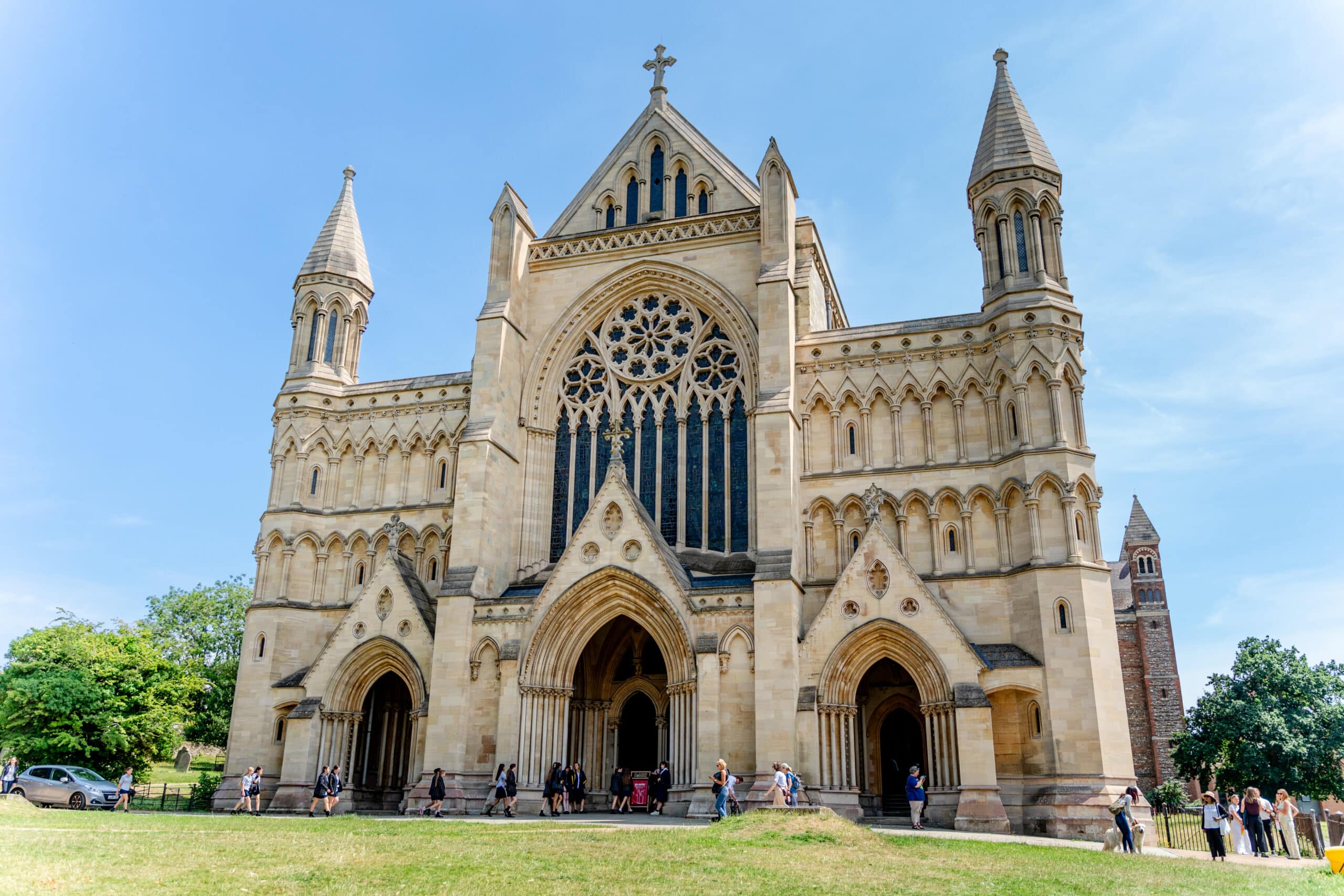 A large, ornate Gothic-style cathedral with tall towers, arched doorways, and a detailed rose window, stands under a blue sky. People are walking near the entrance and across the grassy area in front.