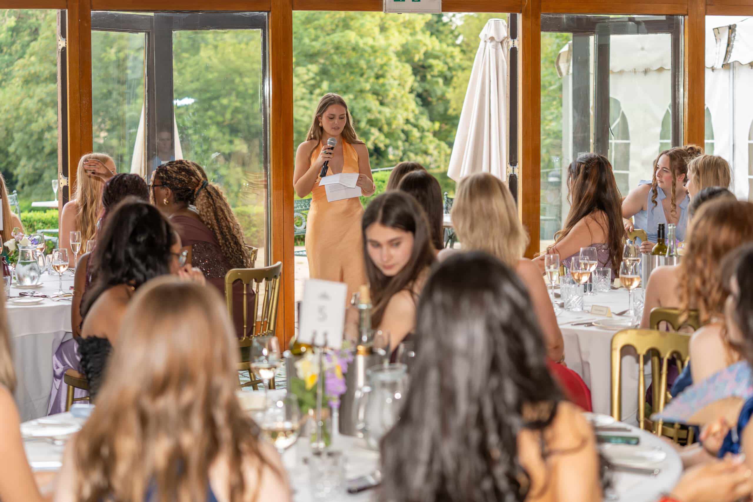 A woman stands and speaks into a microphone in front of large windows, addressing a seated group of people at round tables during a formal indoor event.